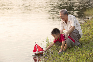 Grandparent helping grandchild with model boat by lake.