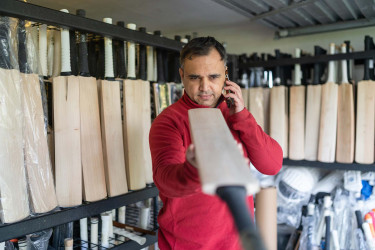 Man talking on his mobile phone with one hand and holding a cricket bat up horizontally with this other hand