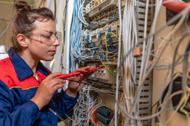 Woman wearing overalls and safety glasses holding a screwdriver and working on an electronics circuit board