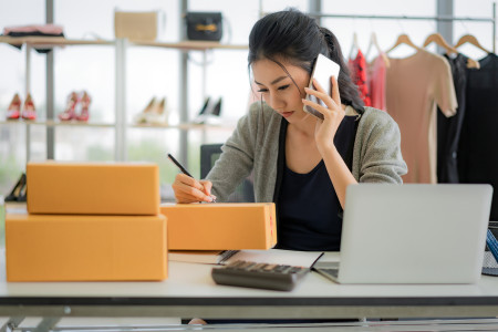 Woman in a fashion shop on talking on her mobile phone while writing on boxes to send out.