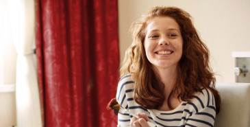 Young person in kitchen eating cake by window and smiling.