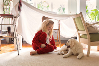 Child sits indoor blanket play house with puppy.