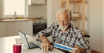 Elderly person in wheelchair at laptop sorting paper documents