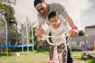 Parent helping toddler ride bike in garden