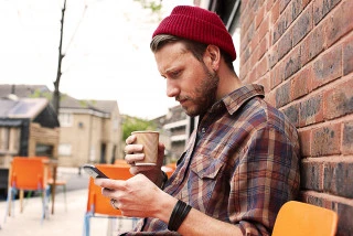 Person leaning against brick wall, using mobile and holding takeaway coffee cup