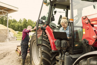Two farm workers, one sitting in tractor using laptop while other stands by