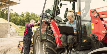 Two farm workers, one sitting in tractor using laptop while other stands by